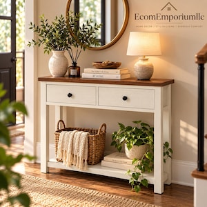 May include: A white console table with a dark wood top, two drawers with black knobs, and a lower shelf. The table holds a vase with greenery, a reed diffuser, a wooden bowl, two books, and a lamp. A woven basket with a cream throw and a plant sit below.