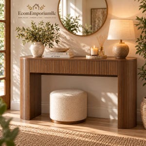 May include: A brown wooden console table with a fluted front design, featuring a rounded edge, paired with a cream-colored textured round ottoman. The table holds a vase with greenery, books titled The Kinfolk Home At Home, and a lamp.