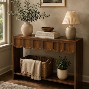 May include: A dark wood console table with a scalloped apron design, featuring a lower shelf holding a woven wicker basket with a beige throw blanket and a potted plant. The top surface displays a vase with greenery, a wooden bowl, and books.