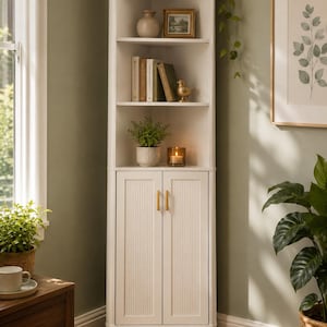 May include: A tall white corner cabinet featuring three open shelves and a lower storage section with two doors. The doors have vertical ribbed detailing and gold-toned handles. The unit is positioned in a corner and holds books, a vase, a small bird figurine, a potted plant, and a candle.