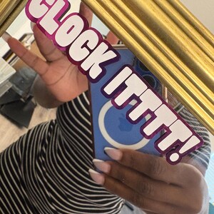 May include: A gold framed mirror with the text "CLOCK ITTTT!" in white letters with a pink outline. The mirror reflects a person wearing a black and white striped top and holding a blue phone.
