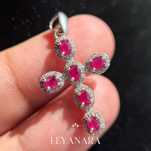 Peut inclure: Pendentif en argent en forme de croix, orné de pierres précieuses rouges vives de taille ovale, chacune entourée d'un halo de petites pierres claires. Le pendentif a une boucle en haut pour une chaîne.