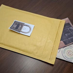 May include: A yellow bubble mailer with a sticker featuring a detailed illustration of a lock. Two additional cards are partially visible, one with a geometric design and the other with concentric circles.