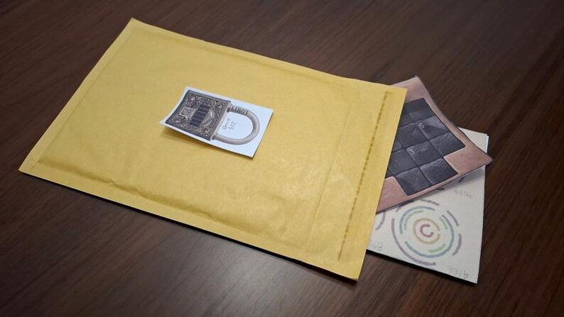 May include: A yellow bubble mailer with a sticker featuring a detailed illustration of a lock. Two additional cards are partially visible, one with a geometric design and the other with concentric circles.