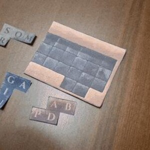 May include: A wooden puzzle with several lettered pieces and a larger rectangular base. The pieces are various shades of gray and blue, with letters printed on them. The base is a light brown color. The letters on the pieces spell out words.