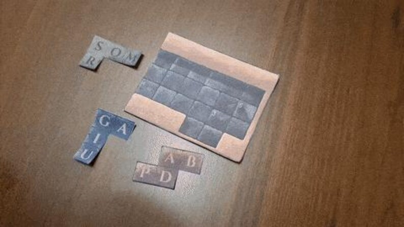 May include: A wooden puzzle with several lettered pieces and a larger rectangular base. The pieces are various shades of gray and blue, with letters printed on them. The base is a light brown color. The letters on the pieces spell out words.