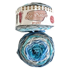 May include: A skein of Lion Brand Ice Cream Deluxe yarn in shades of blue, gray, and white. The yarn is soft and plush, with a gradient effect. The packaging features the words "Ice Cream Deluxe".