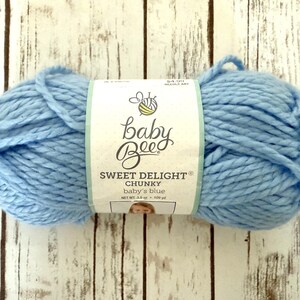 Pode incluir: Um novelo de fio Baby Bee Sweet Delight Chunky em azul bebé. O fio azul claro é embrulhado com um rótulo que mostra o nome da marca e os detalhes do produto. O novelo pesa 100 g.