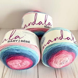 May include: Three balls of Mandala Baby yarn in shades of pink, blue, and teal. Each ball has a label with the word "mandala" in cursive script. The yarn is arranged on a light-colored wooden surface.
