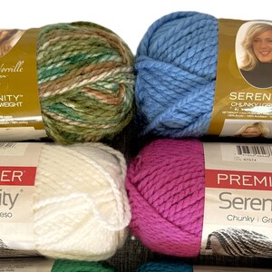 May include: A stack of colorful yarn skeins. The top skein is green and brown, labeled "Deborah Norville Collection Serenity Chunky Weight." Below are a blue skein and a white skein labeled "Premier Serenity Chunky | Grueso." A pink skein is also visible.