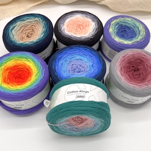 May include: Seven skeins of colorful yarn are arranged on a white surface. The yarn is in various colors, including blue, green, purple, and red. Each skein has a gradient effect, with the colors blending into each other. The yarn is labeled "Cotton Kings".