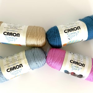 Può includere: Quattro gomitoli di filato Caron Simply Soft, ciascuno del peso di 170 g. I colori includono un beige chiaro, un blu ottanio profondo, un azzurro polvere tenue e un rosa vivace. Ogni gomitolo presenta un'etichetta bianca con il marchio.