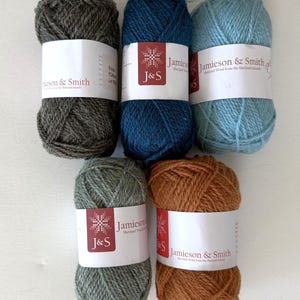 May include: Five skeins of Jamieson & Smith Shetland wool yarn arranged in a group. The colours include dark charcoal, deep navy blue, light sky blue, sage green, and warm copper brown. Each skein features a white paper label with red branding.