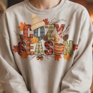 Cozy Season Png with plaid letters and rustic fall details including leaves, pinecones, acorns, and a steaming mug. Features groovy fall retro style with cozy sweater weather accents. Isolated on a white background, perfect for autumn sublimation projects, fall shirts, mugs, crafts, and diy seasonal decorations.