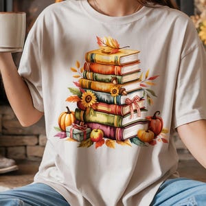 Puede incluir: Camiseta beige con una ilustración de acuarela de una pila de libros con decoraciones otoñales. El diseño incluye calabazas, hojas y un girasol. La camiseta se combina con unos vaqueros azules.