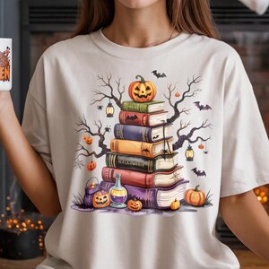 May include: Cream-colored t-shirt featuring a Halloween-themed design. The graphic depicts a stack of books topped with a carved pumpkin, surrounded by bats, spiders, and spooky trees. A matching mug with the words "Happy Halloween" is held in the hand.