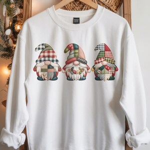 Könnte beinhalten: Weißes Sweatshirt mit drei Zwergen-Illustrationen. Jeder Zwerg hat einen Hut mit unterschiedlichem Karomuster und Patchwork-Kleidung in Rot, Grün und Blau. Die Zwerge haben weiße Bärte und Blumenschmuck.