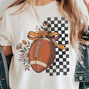 May include: Cream-colored t-shirt featuring a football design. The football is brown with white stitching and a yellow bow. Floral elements and a black and white checkered pattern complete the design.