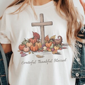 May include: Cream-colored t-shirt featuring a wooden cross, pumpkins, leaves, and a cornucopia. The words "Grateful Thankful Blessed" are printed below the design. The shirt is part of a fall or Thanksgiving theme.