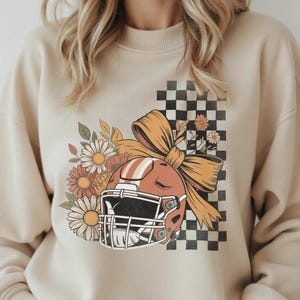 May include: A beige sweatshirt featuring a football helmet design with a bow, flowers, and a checkered background. The helmet is orange and white, with a black face mask. The flowers are in shades of orange, yellow, and white.