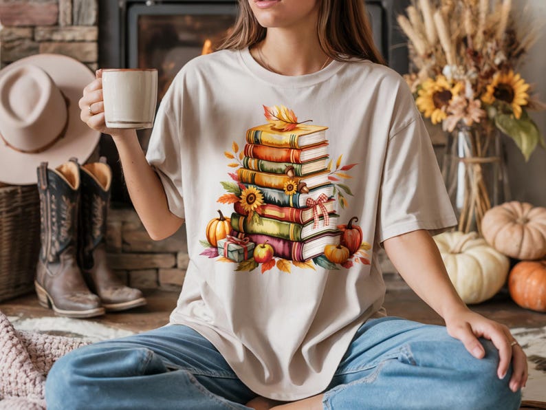 May include: Beige t-shirt featuring a watercolor illustration of a stack of books with fall-themed decorations. The design includes pumpkins, leaves, and a sunflower. The shirt is paired with blue jeans.
