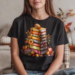 May include: Black t-shirt featuring a watercolor illustration of a stack of books, pumpkins, sunflowers, and fall foliage. The design has a warm color palette of orange, yellow, and red, evoking a cozy autumn theme.