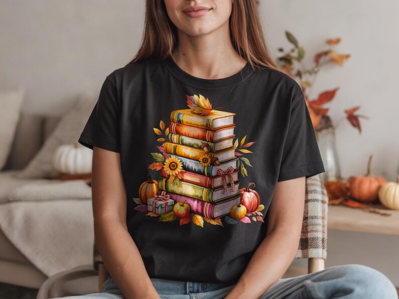 May include: Black t-shirt featuring a watercolor illustration of a stack of books, pumpkins, sunflowers, and fall foliage. The design has a warm color palette of orange, yellow, and red, evoking a cozy autumn theme.