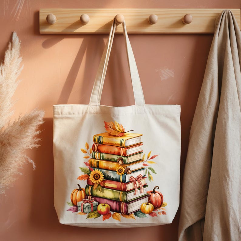 May include: A cream-colored tote bag featuring a stack of books with autumn-themed illustrations. The design includes pumpkins, sunflowers, and fall leaves. The bag has long handles and is hanging on a wooden rack.