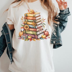 May include: Cream-colored t-shirt featuring a watercolor illustration of a stack of books, pumpkins, sunflowers, and fall foliage. The design includes a gift box and an apple, evoking an autumn theme.