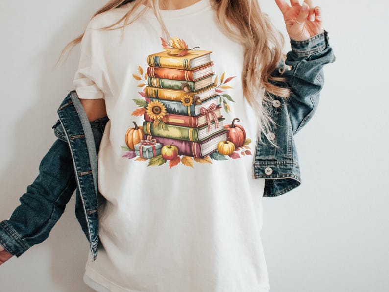 May include: Cream-colored t-shirt featuring a watercolor illustration of a stack of books, pumpkins, sunflowers, and fall foliage. The design includes a gift box and an apple, evoking an autumn theme.