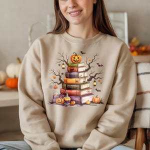 May include: Beige sweatshirt featuring a Halloween-themed graphic. The design includes a stack of books topped with a carved pumpkin, bats, and bare tree branches. Additional pumpkins and lanterns are also present.