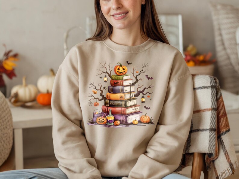 May include: Beige sweatshirt featuring a Halloween-themed graphic. The design includes a stack of books topped with a carved pumpkin, bats, and bare tree branches. Additional pumpkins and lanterns are also present.