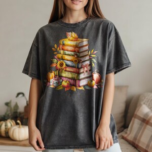 May include: Dark gray t-shirt featuring a watercolor illustration of a stack of books, pumpkins, apples, and fall leaves. The design has a vintage, distressed look, perfect for autumn. The shirt is a comfortable, casual style.