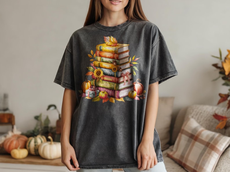 May include: Dark gray t-shirt featuring a watercolor illustration of a stack of books, pumpkins, apples, and fall leaves. The design has a vintage, distressed look, perfect for autumn. The shirt is a comfortable, casual style.