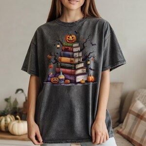 May include: Dark gray t-shirt featuring a Halloween-themed graphic. The design includes a stack of books topped with a jack-o'-lantern, bats, and pumpkins. The shirt has a vintage, washed look.