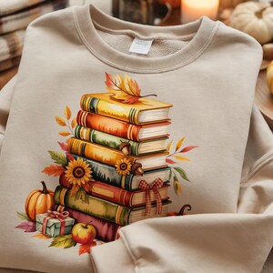 May include: Beige sweatshirt featuring a watercolor illustration of a stack of books with fall-themed decorations. The design includes sunflowers, pumpkins, leaves, and a small gift box, evoking an autumn or harvest theme.