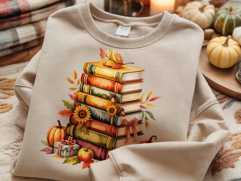 May include: Beige sweatshirt featuring a watercolor illustration of a stack of books with fall-themed decorations. The design includes sunflowers, pumpkins, leaves, and a small gift box, evoking an autumn or harvest theme.