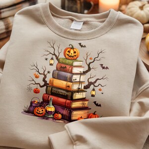 May include: Beige sweatshirt featuring a Halloween-themed graphic. The design includes a stack of books topped with a carved pumpkin, surrounded by bats, spiders, and bare tree branches. Additional pumpkins and a potion bottle are also visible.