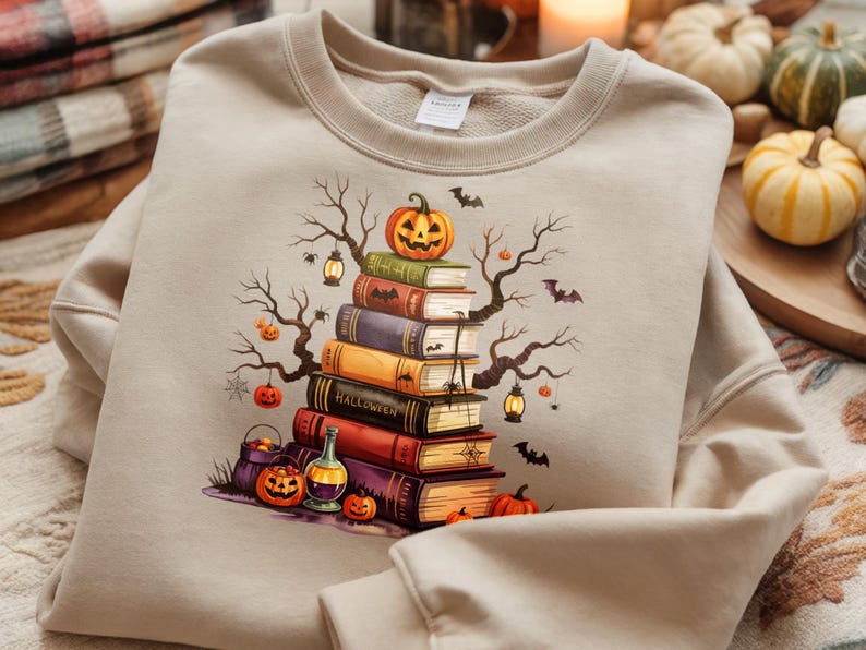 May include: Beige sweatshirt featuring a Halloween-themed graphic. The design includes a stack of books topped with a carved pumpkin, surrounded by bats, spiders, and bare tree branches. Additional pumpkins and a potion bottle are also visible.