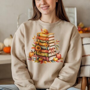 May include: Beige sweatshirt featuring a watercolor illustration of a stack of books, pumpkins, sunflowers, and fall leaves. The design evokes a cozy autumn theme, perfect for book lovers and fall enthusiasts.