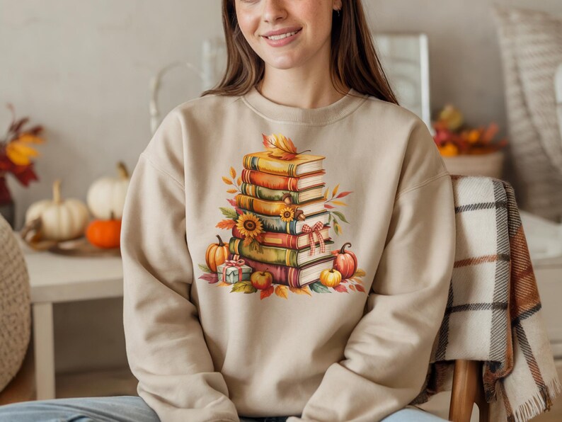 May include: Beige sweatshirt featuring a watercolor illustration of a stack of books, pumpkins, sunflowers, and fall leaves. The design evokes a cozy autumn theme, perfect for book lovers and fall enthusiasts.