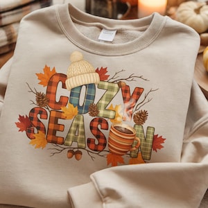 May include: Beige sweatshirt with the words "Cozy Season" in a plaid design, featuring a knit hat, fall leaves, pine cones, and a mug of coffee. The sweatshirt is folded on a patterned surface.