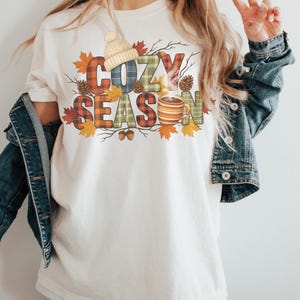 May include: Cream-colored t-shirt with the words "COZY SEASON" in plaid letters, decorated with fall leaves, acorns, and a knit hat. A steaming mug of coffee is also featured in the design. The shirt is worn with a denim jacket.