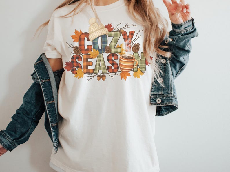 May include: Cream-colored t-shirt with the words "COZY SEASON" in plaid letters, decorated with fall leaves, acorns, and a knit hat. A steaming mug of coffee is also featured in the design. The shirt is worn with a denim jacket.