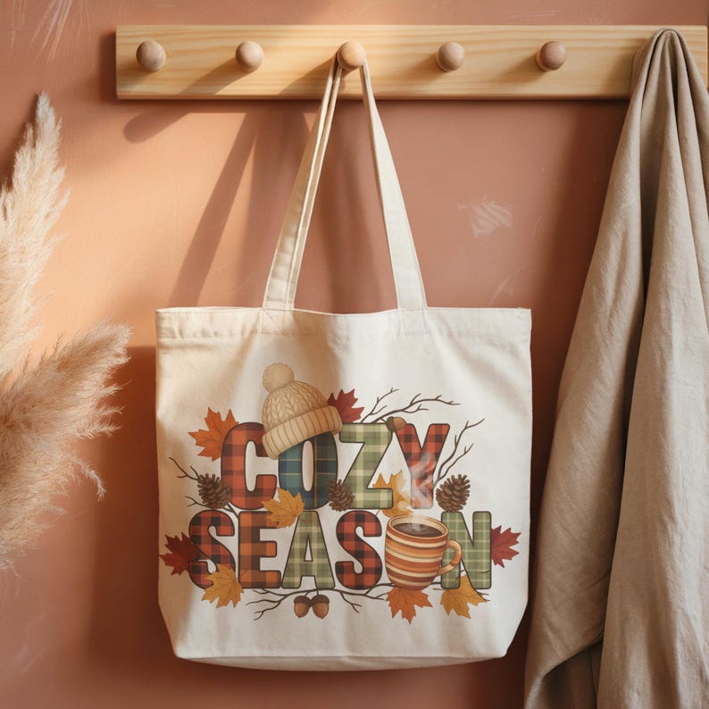 May include: Cream-colored tote bag with the words "COZY SEASON" in a fall-themed design. The design includes a knit hat, coffee cup, autumn leaves, and pine cones. The bag is hanging on a wooden rack.
