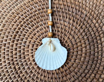 May include: A white seashell pendant with a small shell accent, strung on a natural cord with wooden beads. The pendant hangs against a woven, brown background, creating a coastal or beach-themed aesthetic.
