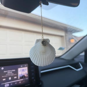 May include: A white seashell car charm hanging from a gold chain. The seashell has a detailed, ridged texture and is adorned with a small gold charm. The charm is hanging from the rearview mirror.