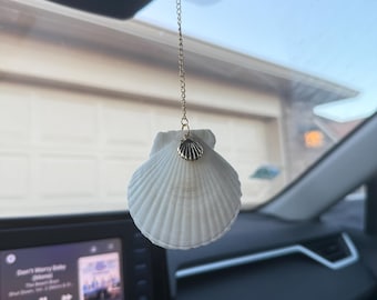 May include: A white seashell car charm hanging from a gold chain. The seashell has a detailed, ridged texture and is adorned with a small gold charm. The charm is hanging from the rearview mirror.