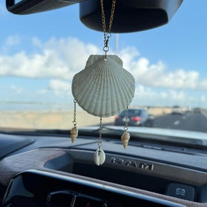 May include: A hanging car ornament featuring a large, white, fan-shaped scallop shell suspended by a gold-toned chain. Three smaller, distinct seashells hang from the bottom of the main shell on individual gold-toned chains, creating a mobile effect.