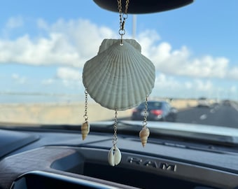 May include: A hanging car ornament featuring a large, white, fan-shaped scallop shell suspended by a gold-toned chain. Three smaller, distinct seashells hang from the bottom of the main shell on individual gold-toned chains, creating a mobile effect.